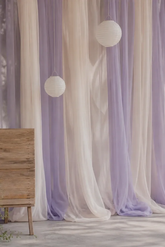 A photo backdrop made from layered sheer fabric panels in pale lavender and cream, draped in vertical strands. Several small paper lanterns hang at different heights, and a simple wooden sign stands in front.