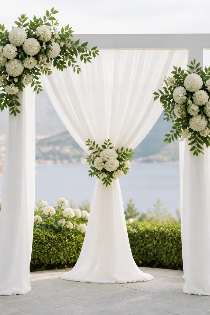 A white wedding arch draped with sheer fabric panels that hang in soft folds. Matte white round flower clusters sit near the top and center, with green leaves creating depth.