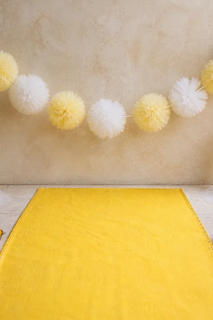 A simple yellow runner on the floor with a garland of white and pale yellow tulle pom-poms strung above, lightly touching the runner near the sides; marigold accents sit at the tray corners.