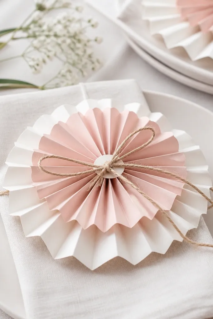 White and blush paper rosettes stacked like flowers around folded napkins; small script labels are tied on with twine.