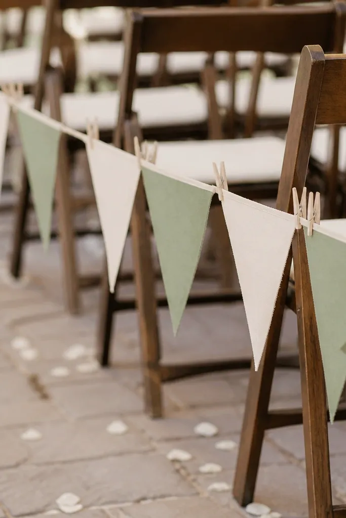 Small triangular fabric flags in cream and sage on a string, clipped with wooden clothespins to a thin line across chairs along the aisle.