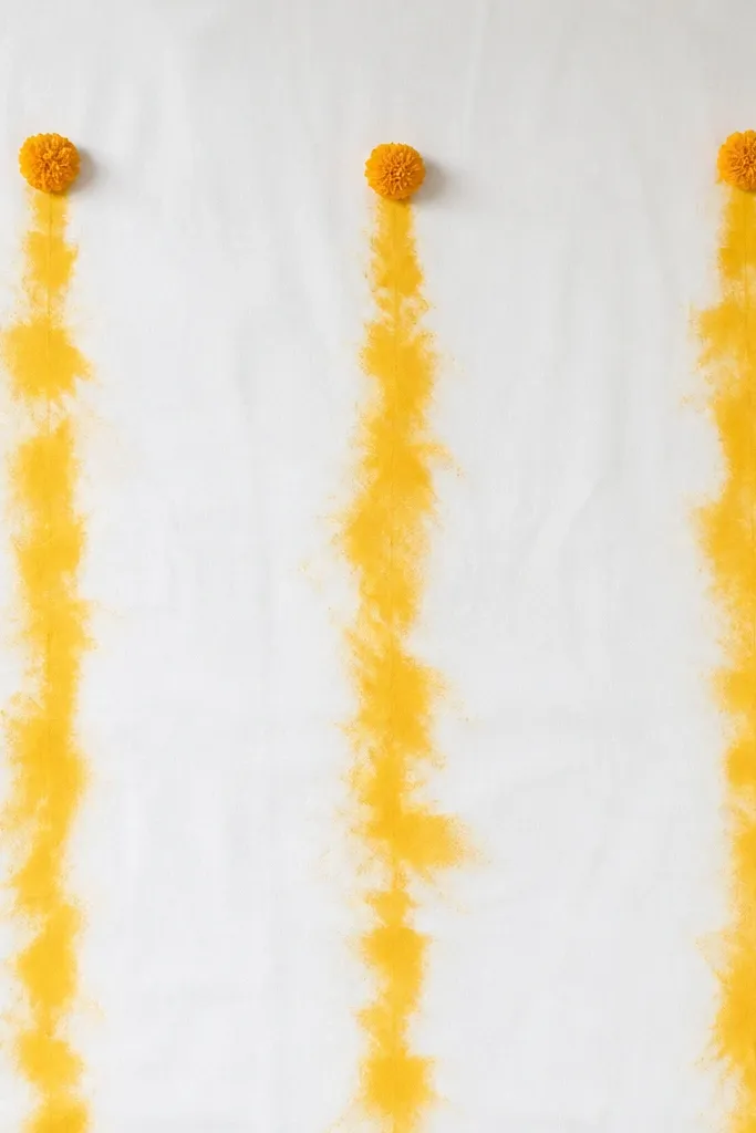 A plain white sheet backdrop with three vertical turmeric-yellow sash lines. The lines look like soft tie-dye smudges, and tiny marigold buds sit at the top of each sash line.