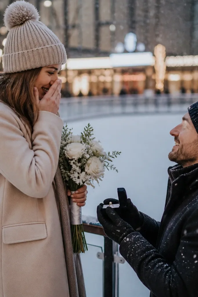 A person holds a small bouquet while standing just inside an outdoor ice rink boundary. Snowflakes fall lightly, and the background shows blurred lights from nearby shops. The ring box is open on a gloved palm, and the couple wear matching beanies.