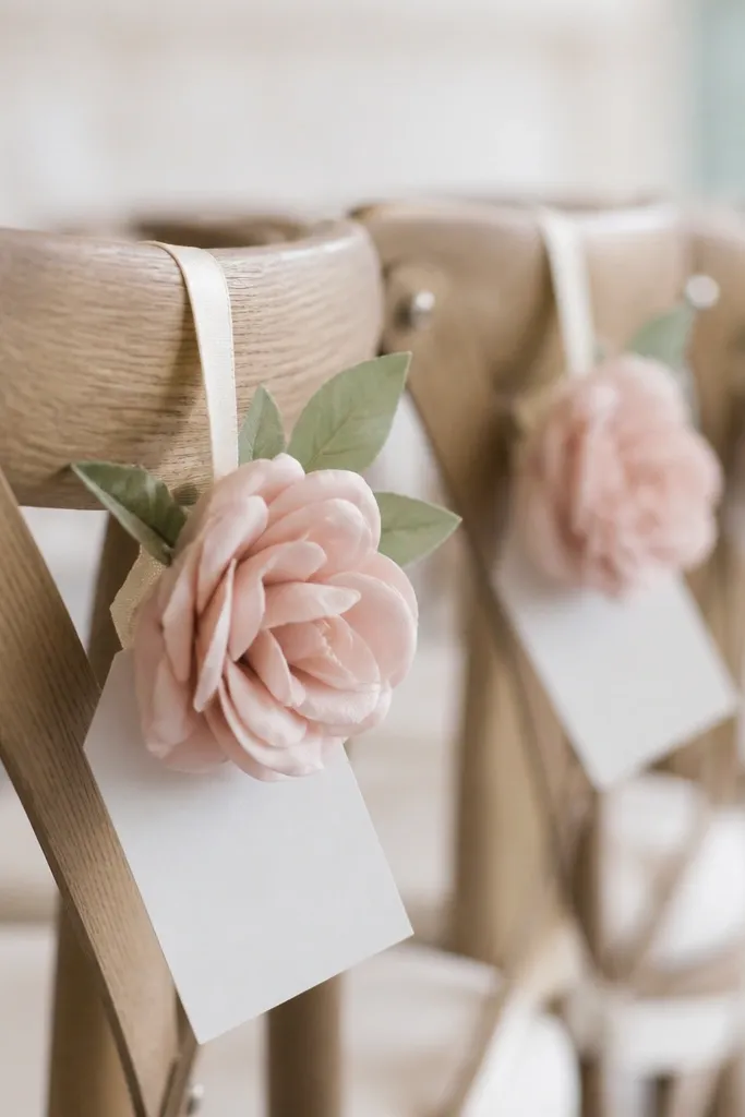 Small seat markers clipped to chair backs with a ribbon loop holding one blush faux flower and a white name tag.