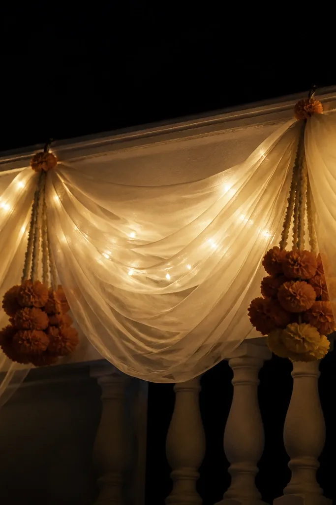 Ivory sheer fabric drapes along a balcony rail with warm gold fairy lights woven in. Small marigold clusters hang from two points. The background is night-dark, and the lights glow softly.