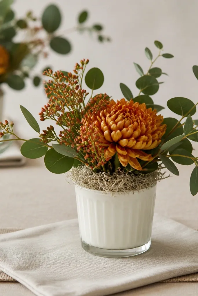 Short milk glass vases with burnt orange blooms, dark green eucalyptus leaves fanning outward, and a base of oat-colored moss. Each vase sits on a square of cream linen with a thin burlap strip underneath.