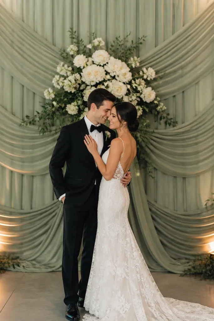 Sage green draped fabric panels fall in waterfall layers behind the couple, with a concentrated cluster of white roses centered at eye level. Soft greenery fills gaps and warm uplights hit the lower drape.