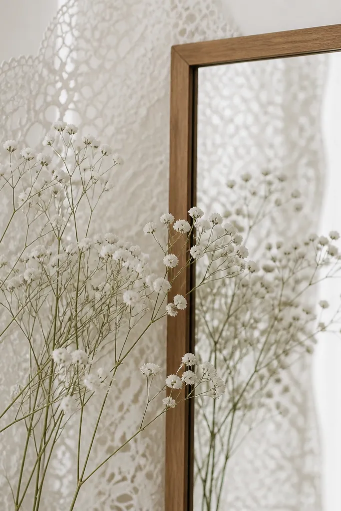 A mirror with a simple wood frame. Behind it, a soft white paper lace sheet covers part of the wall. In front of the lace, several faux baby's breath stems are arranged in a loose fan, and the mirror reflects the airy texture.