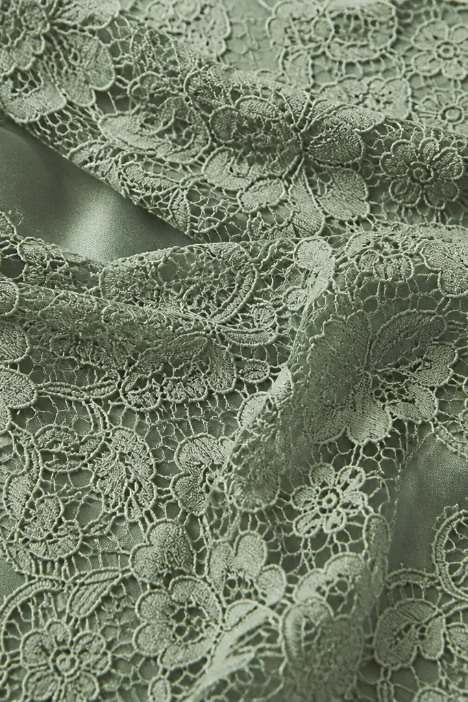 A close-up of sage lace overlay over a smooth satin lining; the lace pattern is crisp and the underlayer shows through softly without patchy transparency.