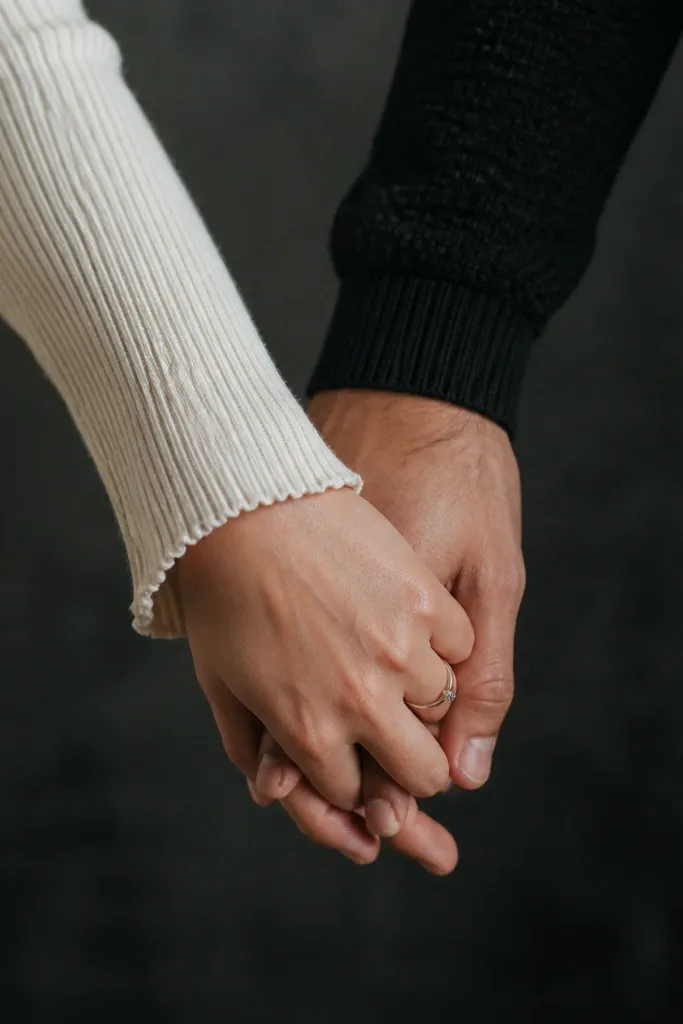 A tight crop shows hands and forearms in the foreground. She wears an ivory ribbed knit sleeve with a delicate cuff, and he wears a black textured sweater. Their fingers interlace at center frame, with a blurred background of a dark studio wall. A small silver ring catches light near the knuckle.