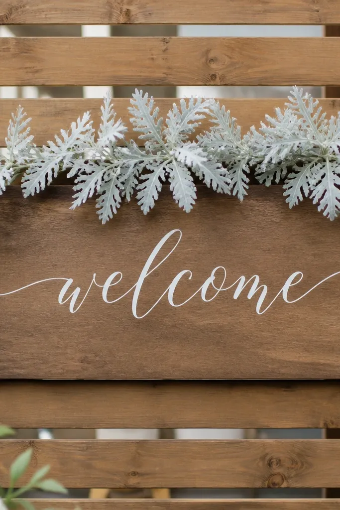 A welcome sign attached to a simple wood slat backdrop. Dusty miller stems spread lightly across the top edge, mostly in pale gray. White lettering sits centered and clean, with no heavy florals.