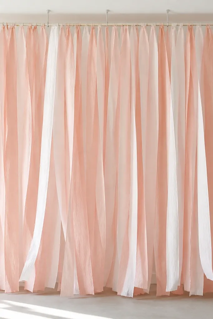 A curtain-style backdrop made of layered blush and white crepe paper strips hanging from a straight rod, attached with small removable ceiling clips; the strips sway slightly.