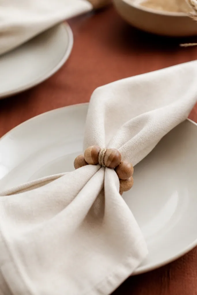 Napkins folded and held by rings made from wooden beads. The beads are grouped into a chunky circle, tied with twine so the ring sits snug. The napkins are cream linen with a rust table runner.