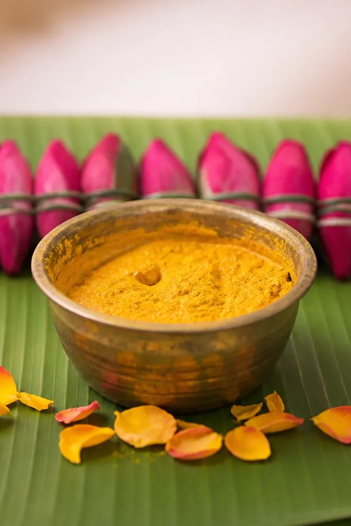 A banana leaf base holds the haldi bowl. Behind the bowl is a straight line of small rose buds tied together, forming a thin backdrop strip. A few petals are scattered at the front base of the bowl.