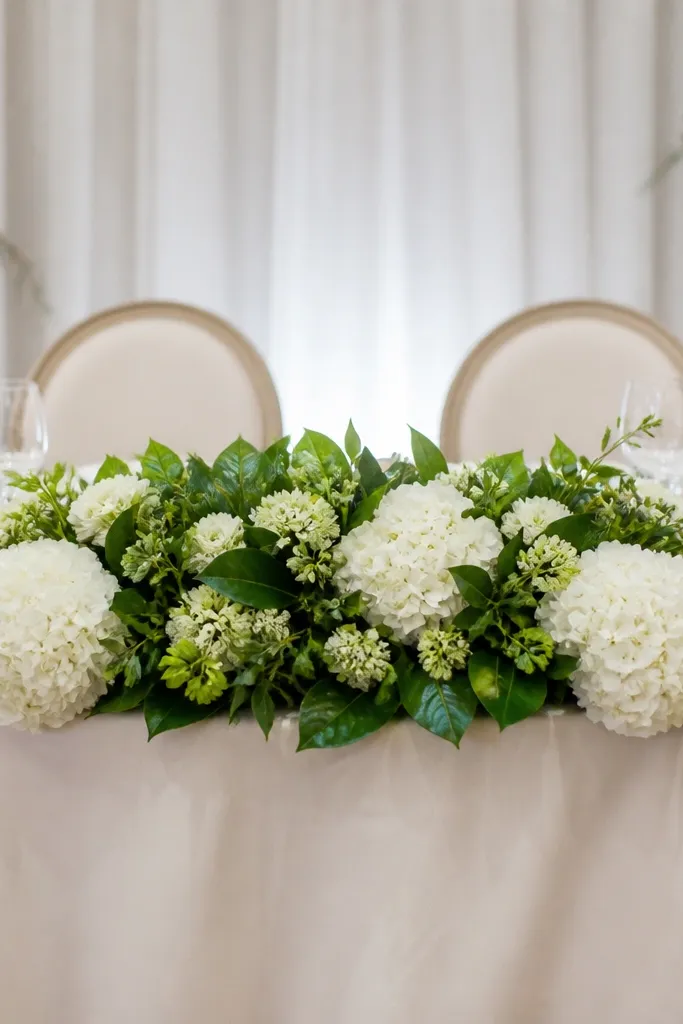 A sweetheart table arrangement with large white hydrangea blooms, glossy green apple-like foliage, and small green buds tucked between. The arrangement is wide and flat, with hydrangea clusters at both front corners.