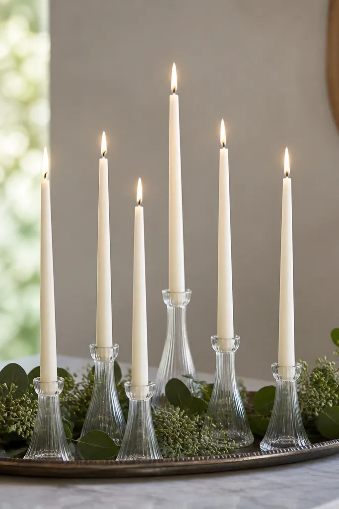 Tapered candles in clear glass holders grouped on a tray with greenery; the candles are slim and evenly spaced.
